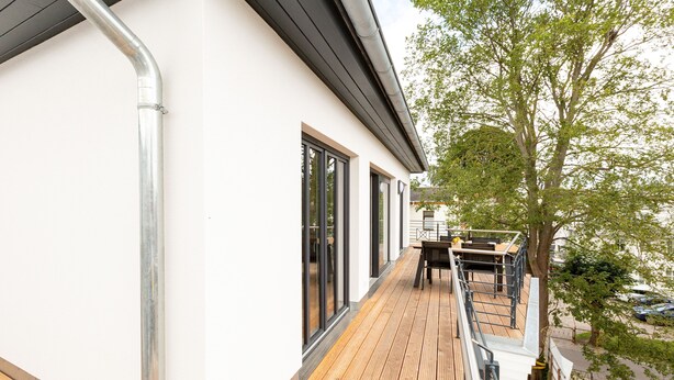 A modern building with a wooden deck, large windows, and a tree in the background.
