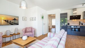 Ein modernes Wohnzimmer mit einer Couch, einem Couchtisch und einem Essbereich. Es gibt eine Küche mit dunklen Schränken und Blick ins Freie.