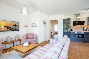 Una sala moderna con sofá, una mesa de centro y un comedor. Se observa una cocina con armarios oscuros y vistas al exterior.
