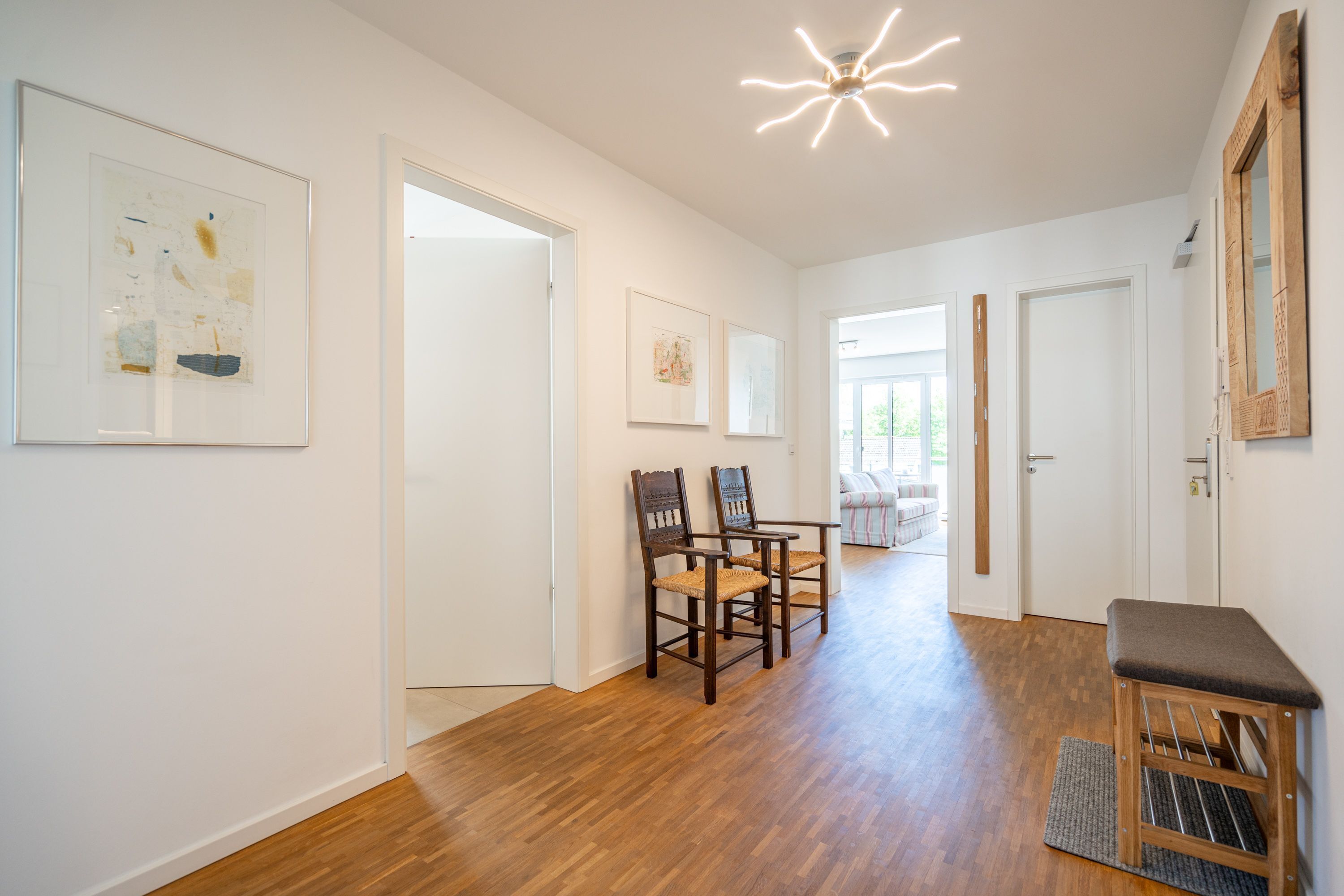 A hallway with wooden flooring, framed artwork on the walls, a bench, and a door leading to another room.