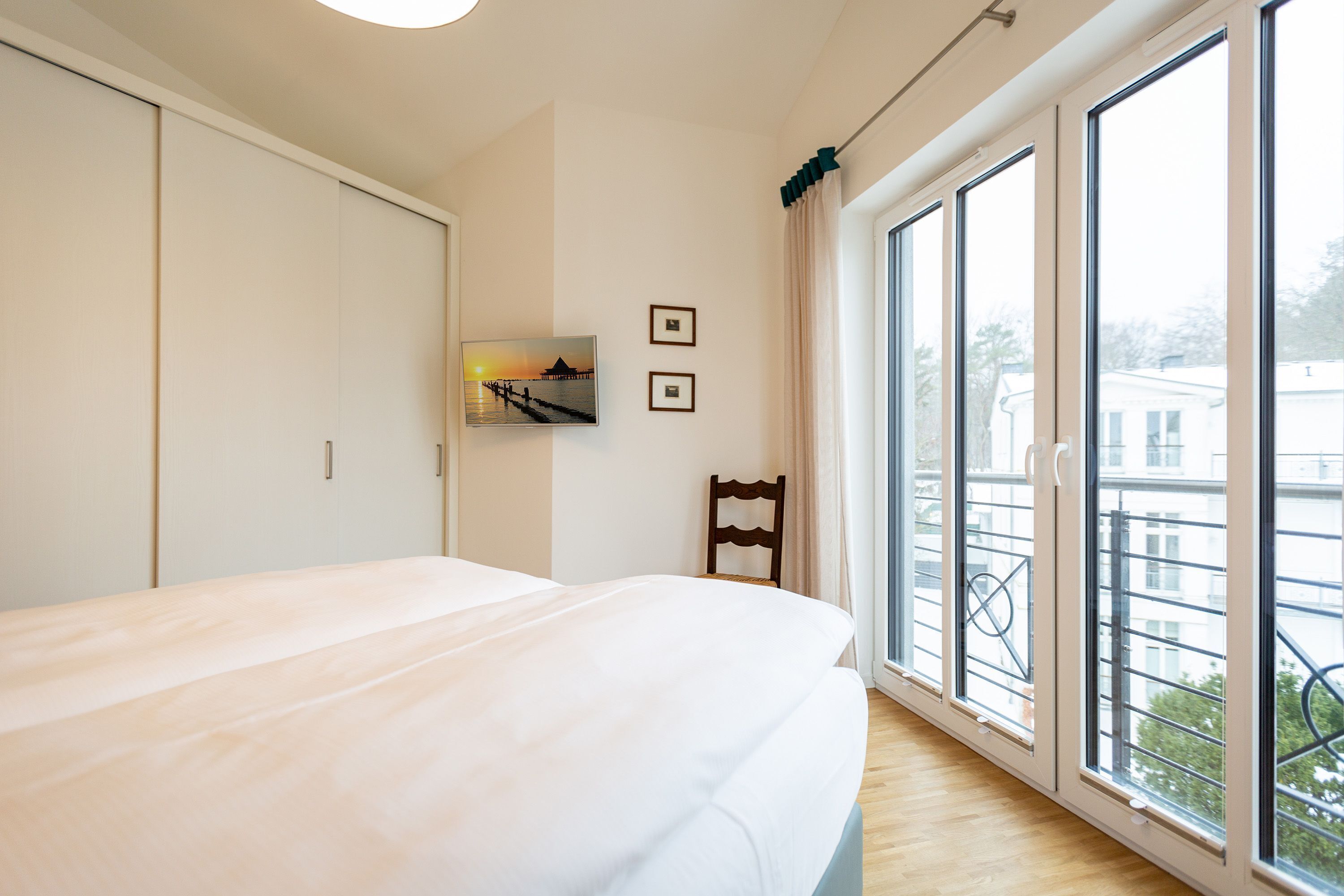 A bedroom with a large bed, a chair, a wall-mounted TV, and a window with a view of buildings.