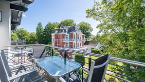 Ein Balkon mit Glastisch und Stühlen, der auf ein rot backsteins Gebäude und üppiges Grün blickt.