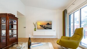 Ein modernes Wohnzimmer mit Holzschrank, gelbem Sessel, Glastisch und einem großen Fernseher an der Wand, der eine Sonnenuntergangsszene zeigt.