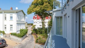 Eine schmale Straße mit einem parkenden Auto am Rand, ein Baum im Vordergrund und Gebäude mit Balkonen und großen Fenstern im Hintergrund.