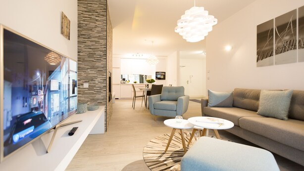 Uma sala moderna com televisão de tela plana, um sofá, duas poltronas, uma mesa de centro e um espaço de jantar ao fundo.