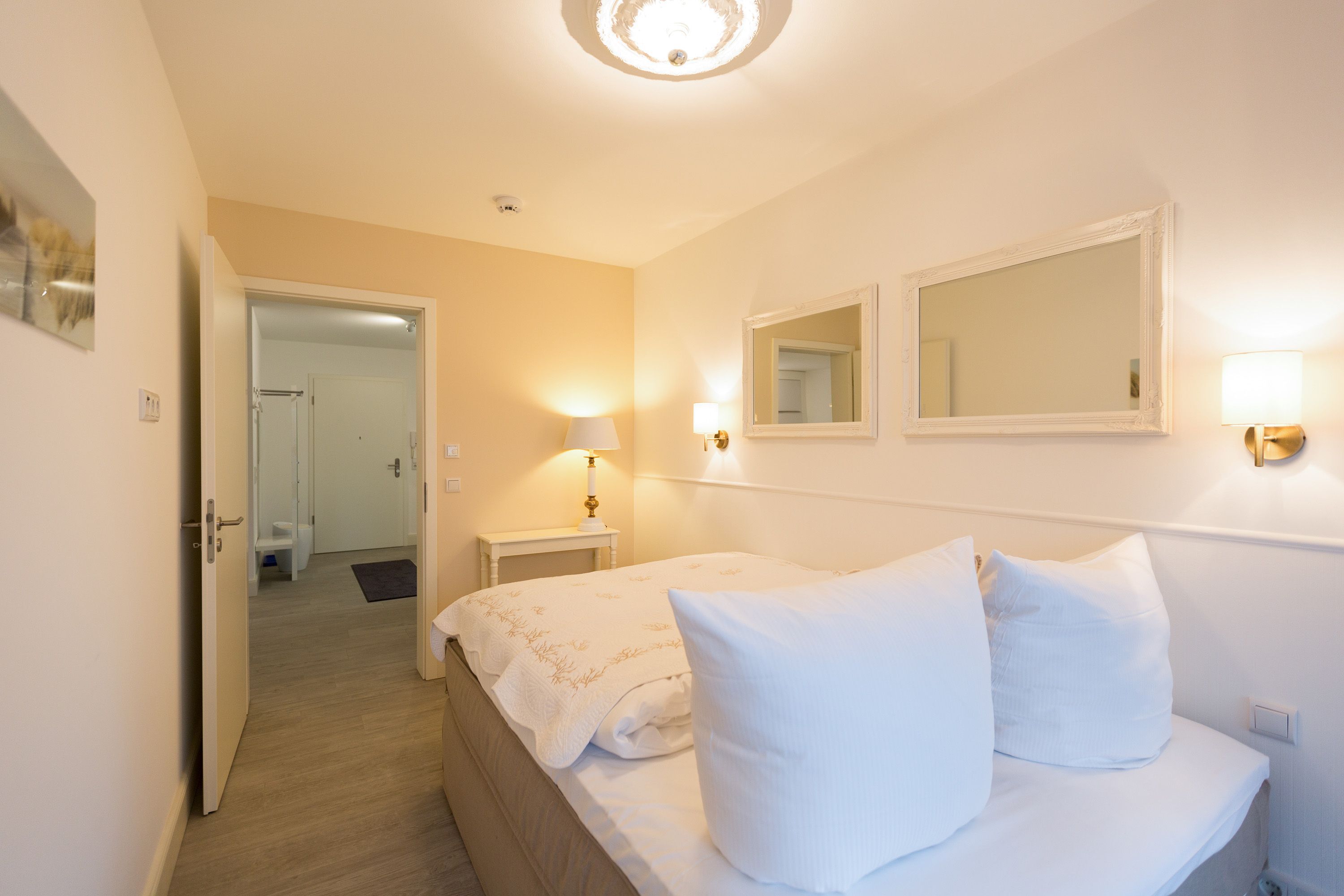 Habitación de hotel con cama, lámparas de noche, un espejo y una puerta que da a un baño.