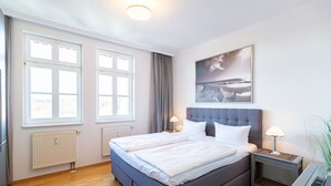 Una camera d'albergo moderna con un letto grande, due comodini, un tavolino, un termosifone e un'ampia finestra con tende.
