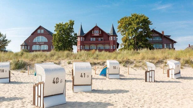 Una fila di cabine sulla spiaggia con numeri, un grande edificio rosso con un cartello 'Vineta' sullo sfondo e un cielo azzurro limpido.
