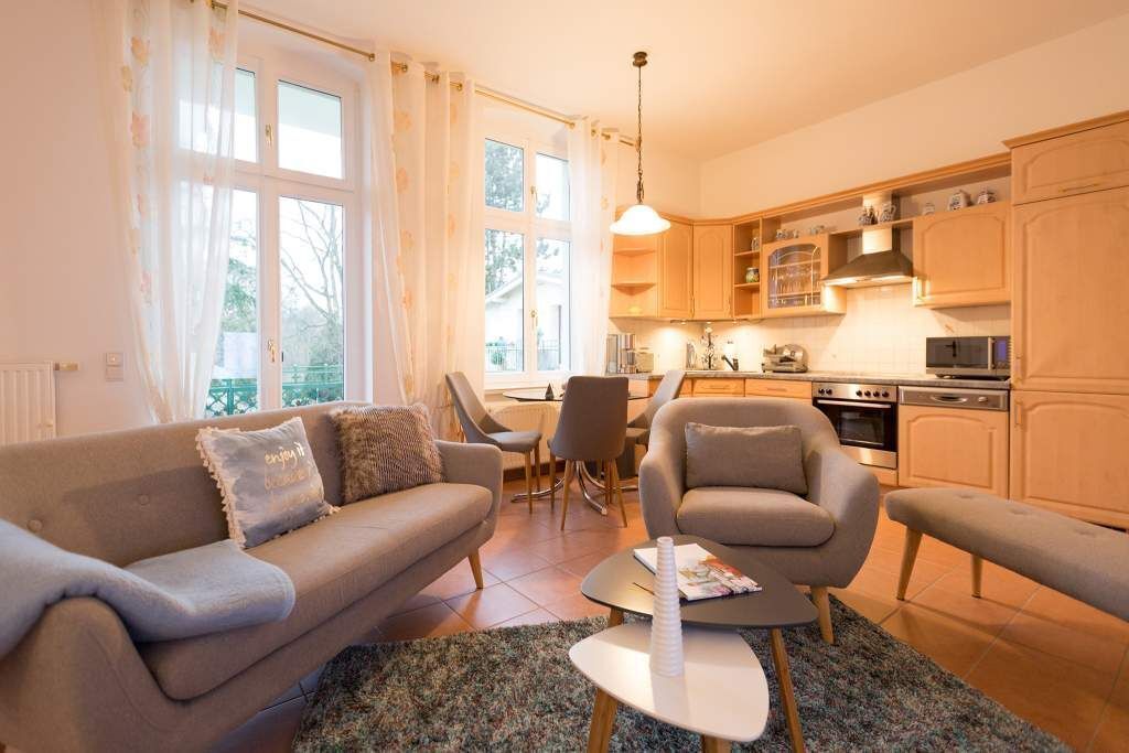 Un salon chaleureux avec un canapé, un fauteuil et une table basse, une cuisine dotée de meubles en bois et d’appareils électroménagers, et de grandes fenêtres avec des rideaux.