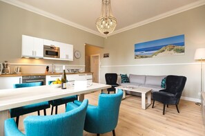 A modern kitchen with white cabinets, a dining area with blue chairs, and a living room with a gray sofa and black armchairs.
