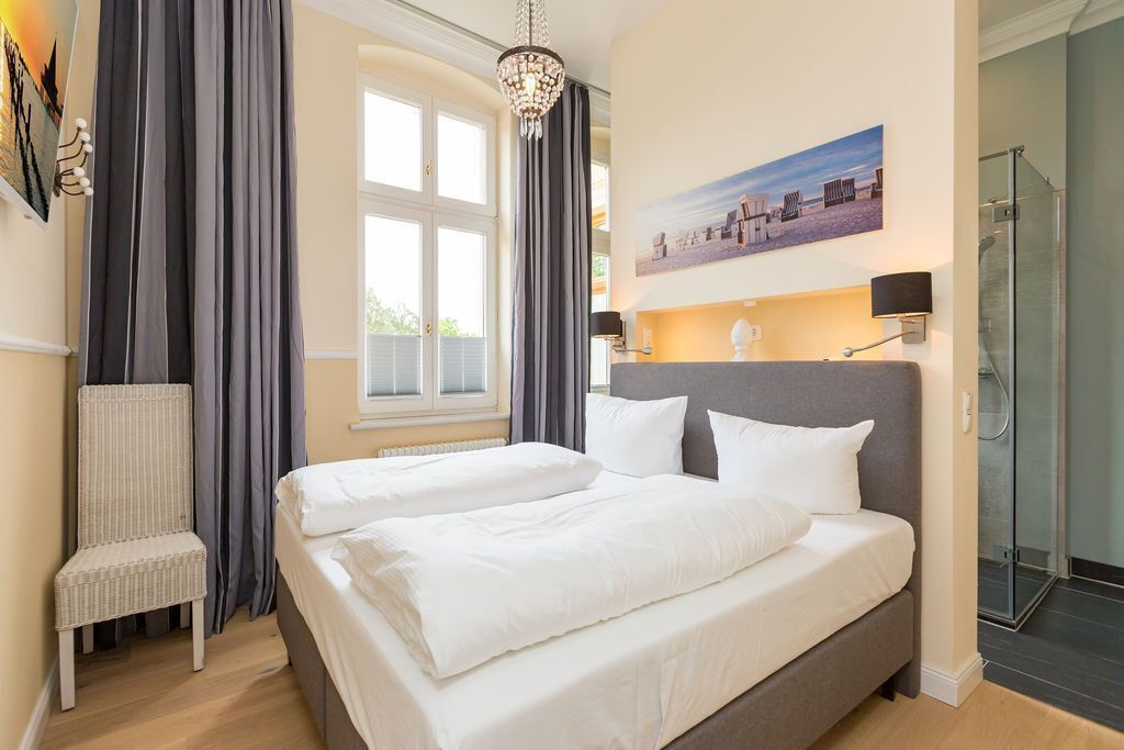 Un lit double avec du linge de lit blanc, une chaise, une grande fenêtre avec des rideaux, un tableau au mur et une douche en verre.