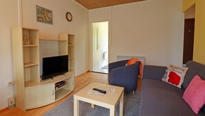 Een woonkamer met een flatscreen televisie op een houten standaard, een grijze bank met een rood kussen en een klein tafeltje met een afstandsbediening.