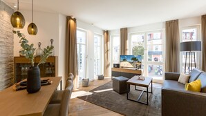Uma sala de estar moderna com uma mesa de jantar de madeira, um sofá, uma mesa de centro e uma televisão. Há uma janela grande com cortinas, uma parede de pedra e uma planta na mesa.