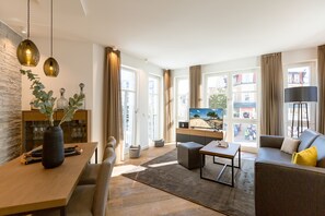 A modern living room with a wooden dining table, a sofa, a coffee table, and a TV. There is a large window with curtains, a stone wall, and a plant on the table. - Beachhouse Bansin, App. 05 (Heringsdorf)