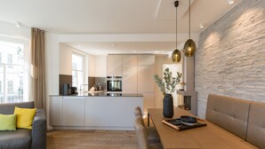 Uma sala de estar moderna com um sofá, uma mesa de jantar com um vaso e tigelas, uma cozinha ao fundo, e uma parede de pedra.