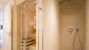 A modern bathroom with a glass-enclosed sauna, a shower area with a handheld showerhead, and a wooden bench.