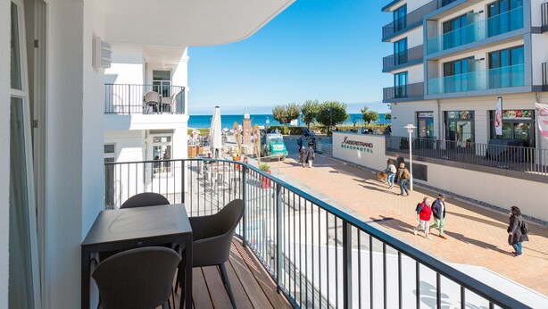 A balcony with a table and chairs, overlooking a beachside promenade with people walking and a beach in the background. - Beachhouse Bansin, app. 04 (Heringsdorf)