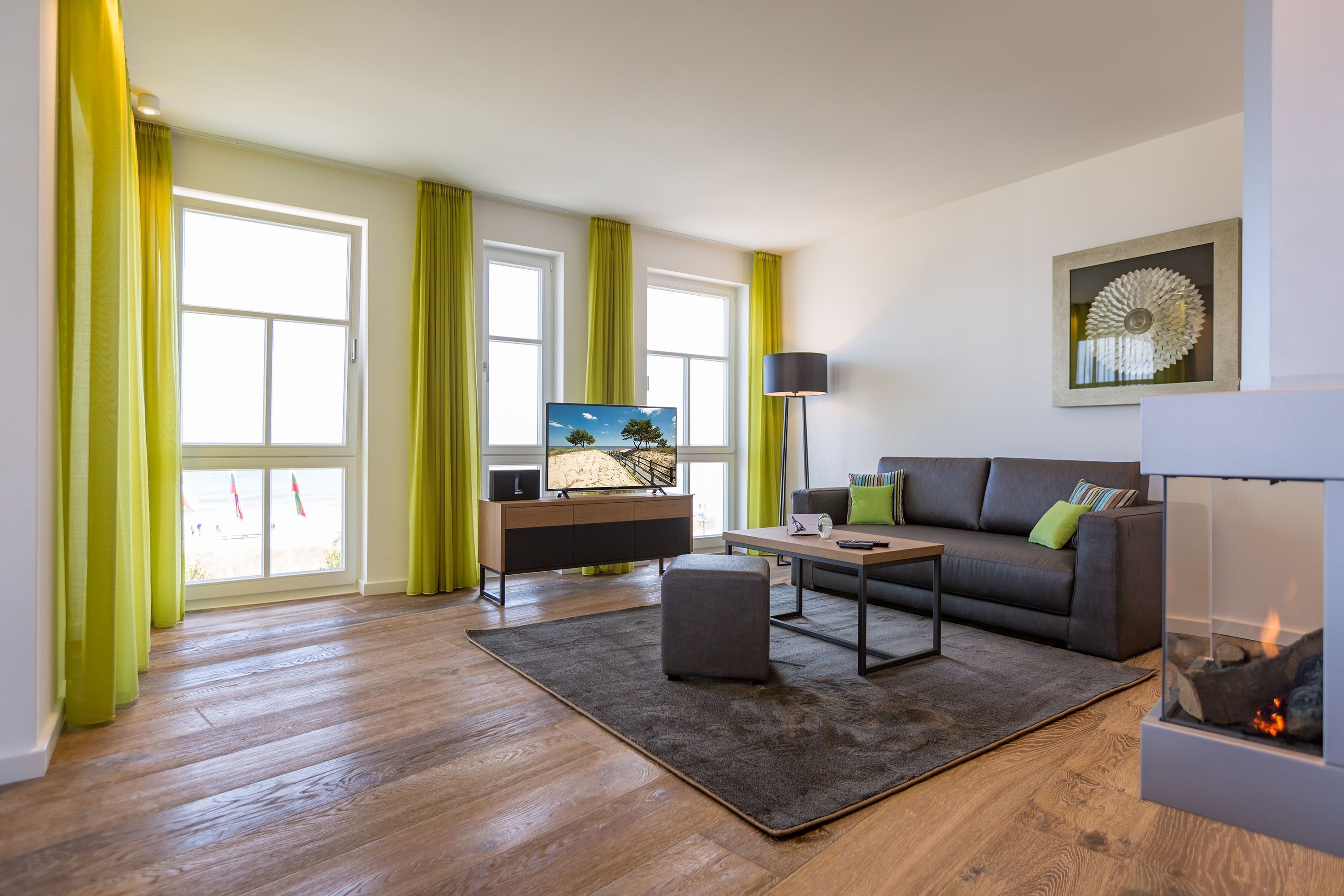 Ein modernes Wohnzimmer mit Sofa, Couchtisch und Fernseher, großen Fenstern mit Vorhängen und einem Kamin.