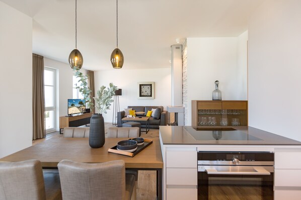 A modern kitchen with a built-in oven, a dining area with a wooden table and chairs, and a living area with a sofa and a TV.