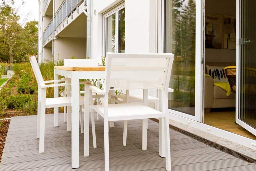 A modern outdoor seating area with white chairs and a wooden table, adjacent to a building with large glass doors.