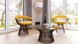 A modern living room with a glass coffee table, two yellow chairs, a floor lamp, and a view of a garden through large windows. - Dune Residence Heringsdorf, App. 05 (Heringsdorf)