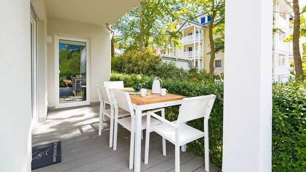 A balcony with a wooden table and white chairs, surrounded by greenery and a building in the background. - Dune Residence Heringsdorf, App. 05 (Heringsdorf)