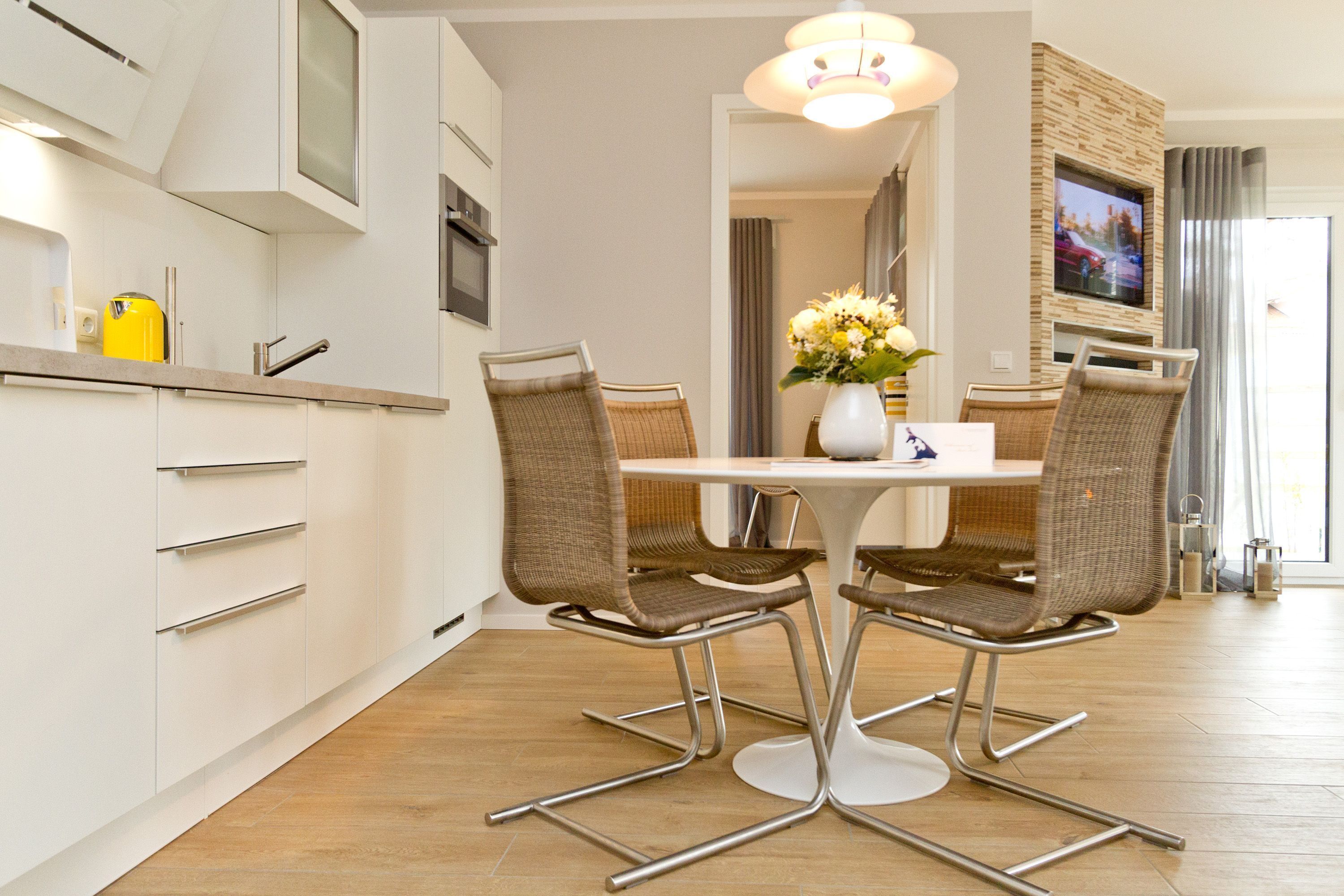 A modern kitchen with white cabinets, a dining table with four wicker chairs, and a TV mounted on the wall.