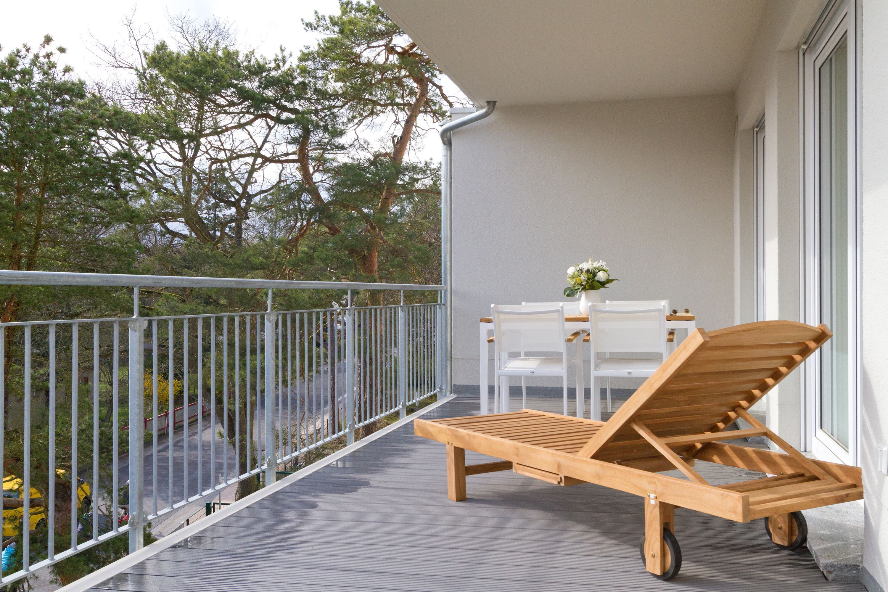 Una poltrona da relax in legno su ruote, un tavolo bianco con sedie e un vaso con fiori bianchi su un balcone con ringhiera in metallo.