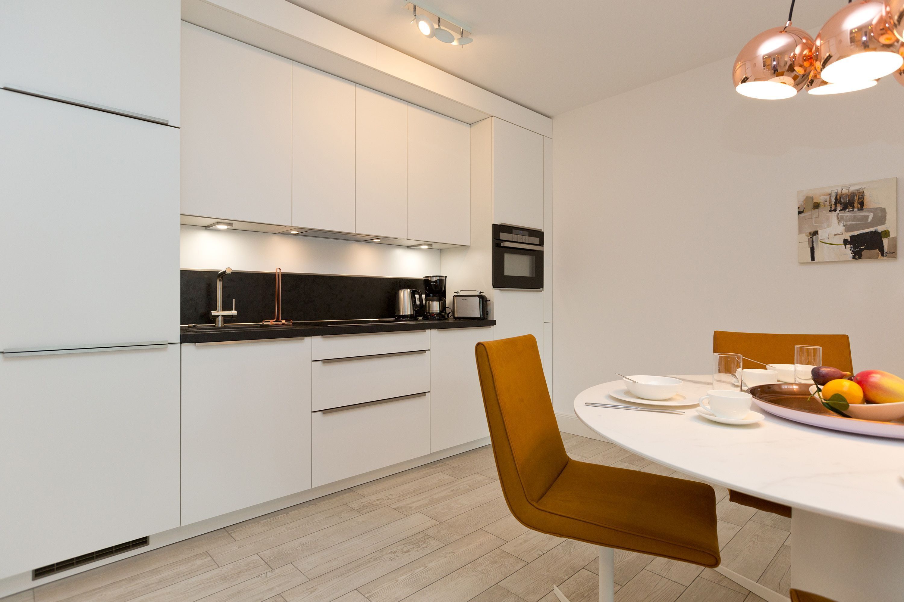 Une cuisine moderne avec des meubles blancs, un plan de travail noir et un coin repas comprenant une table blanche et des chaises marron.