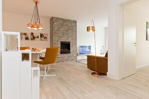 A modern living room with a fireplace, a dining area with a white cabinet and a yellow chair, and a large window.