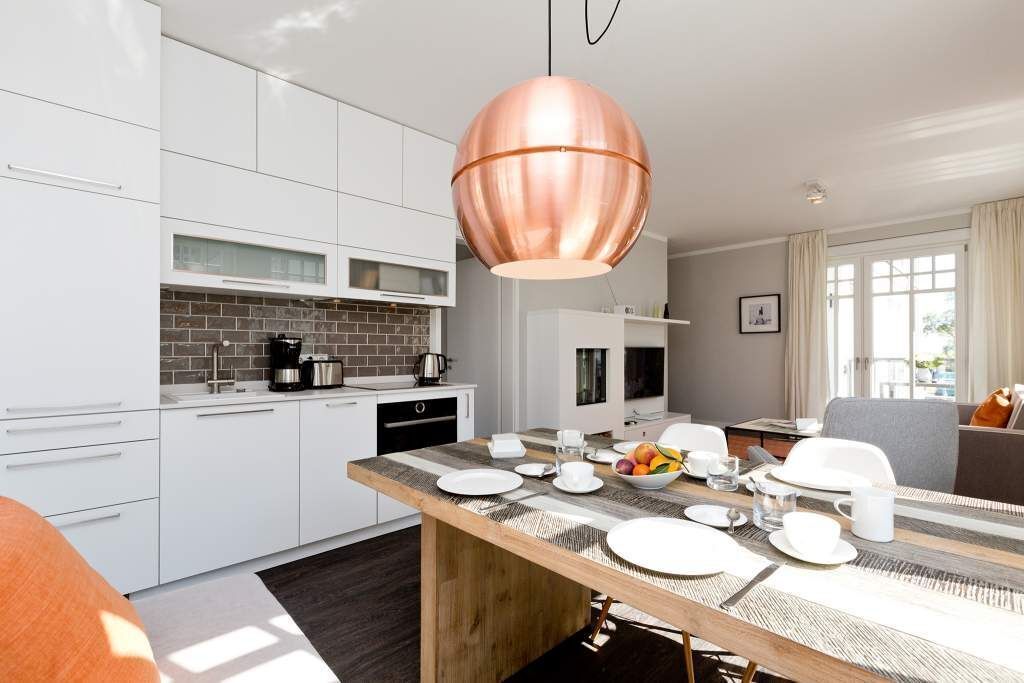 Una cocina moderna con armarios blancos, una lámpara colgante de cobre y un comedor con una mesa de madera dispuesta para cuatro personas.