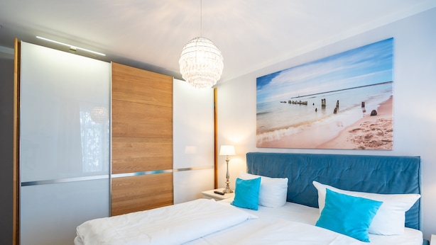 Una cama bien hecha con ropa de cama blanca y azul, un armario de madera, una mesita de noche con una lámpara y una decoración de pared con temática playera.