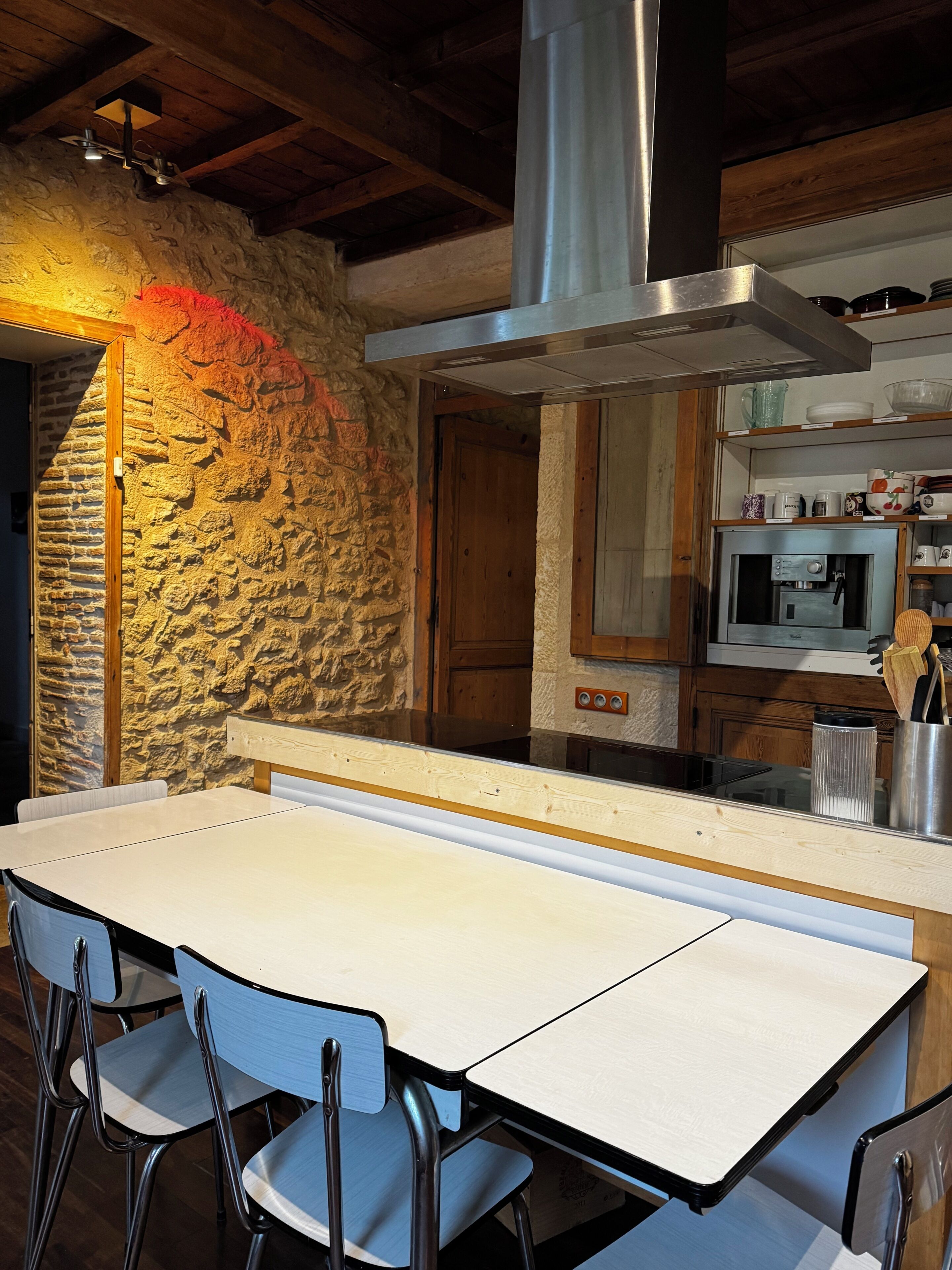 A kitchen with a stone wall, wooden ceiling beams, a stainless steel range hood, and a table with chairs.