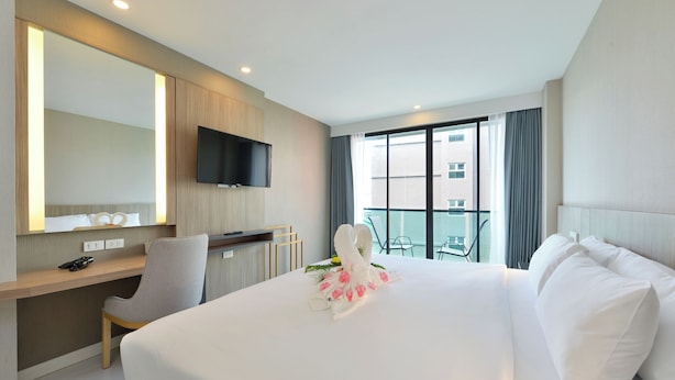 A hotel room with a large bed, a desk with a chair, a flat-screen TV mounted on the wall, and a balcony with a view of the city. - Grace Patong Hotel (Patong)