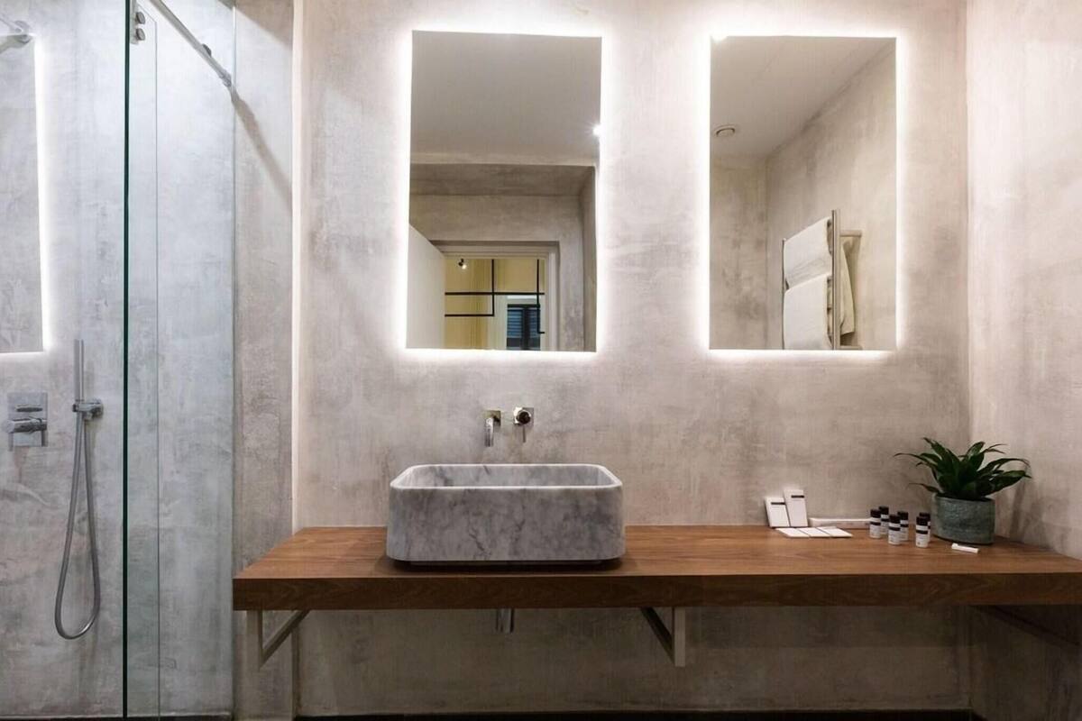 Un baño con lavamanos de mármol, tocador de madera y dos espejos con iluminación integrada.