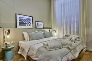 Uma cama bem arrumada com almofadas e um cobertor, uma mesa de cabeceira com abajur, e duas molduras com quadros na parede.