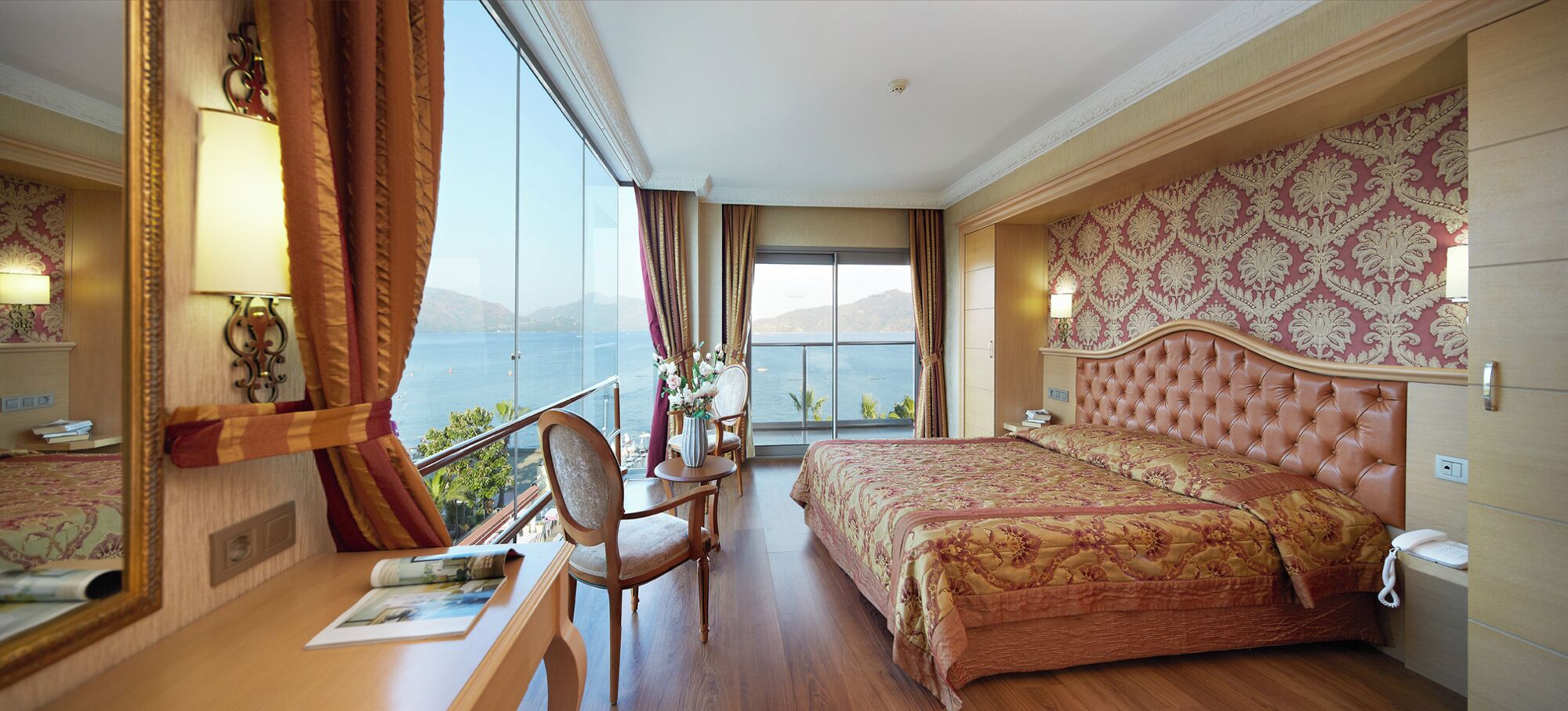 A hotel room with a large bed, a desk with a chair, a view of the sea, and a balcony.