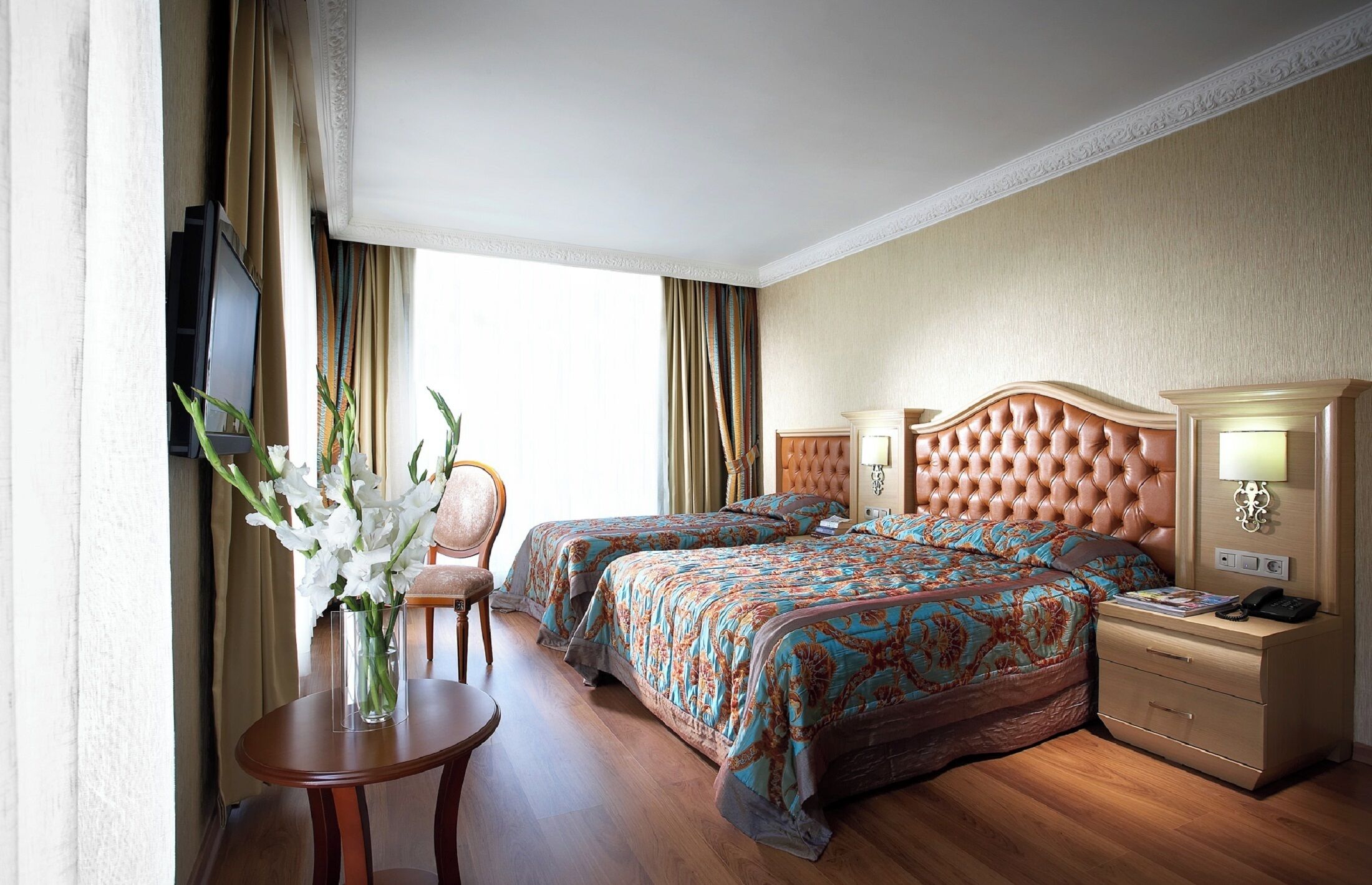 A hotel room with two beds, a bedside table, a chair, a TV, and a vase with flowers.