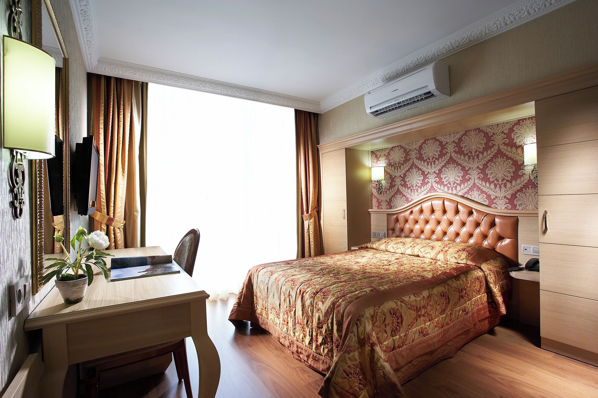 A hotel room with a bed, a desk, a chair, a potted plant, and a wardrobe.