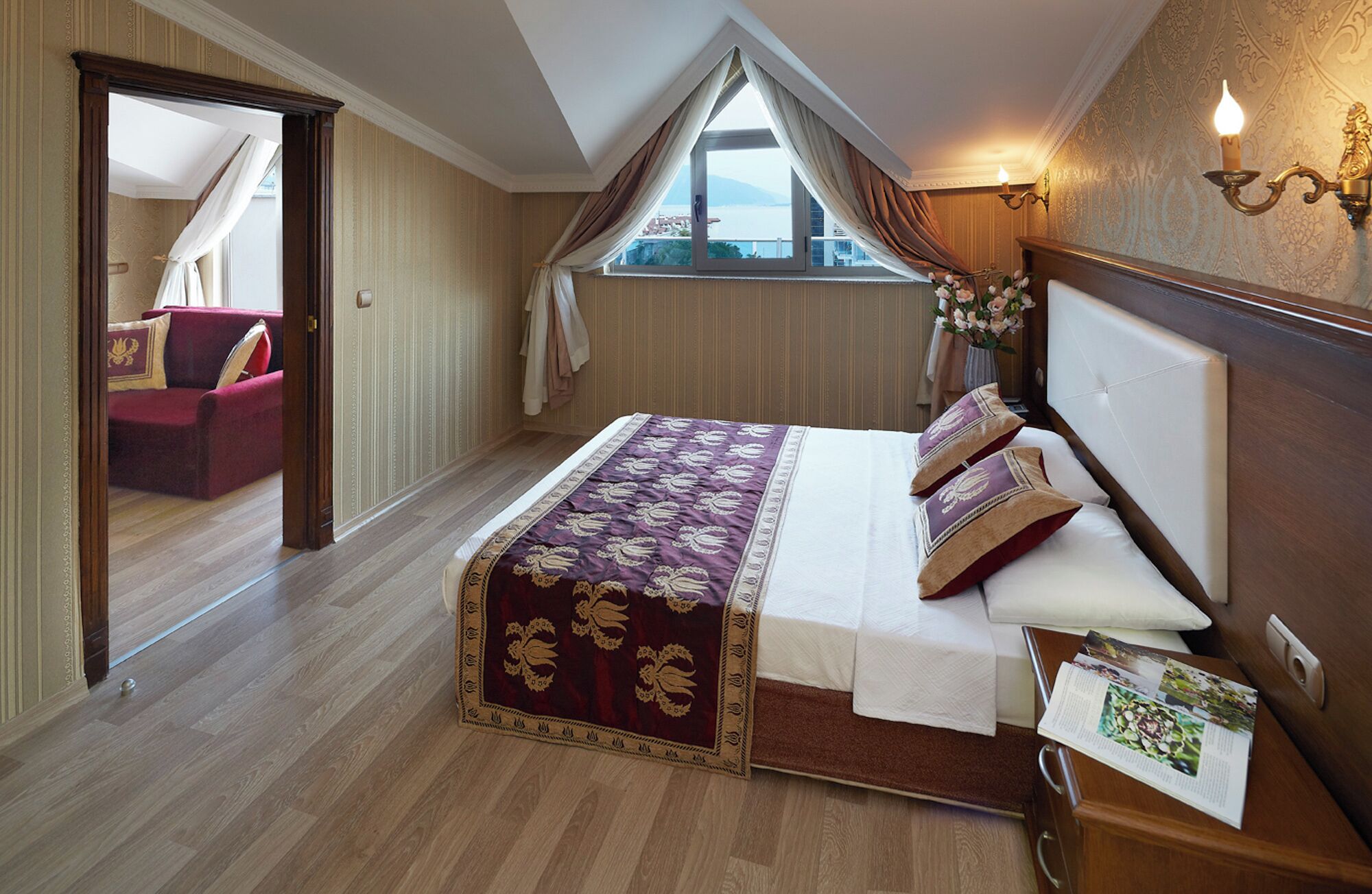 A bedroom with a large bed, a wooden headboard, a window with curtains, a nightstand, and a view of the outside.