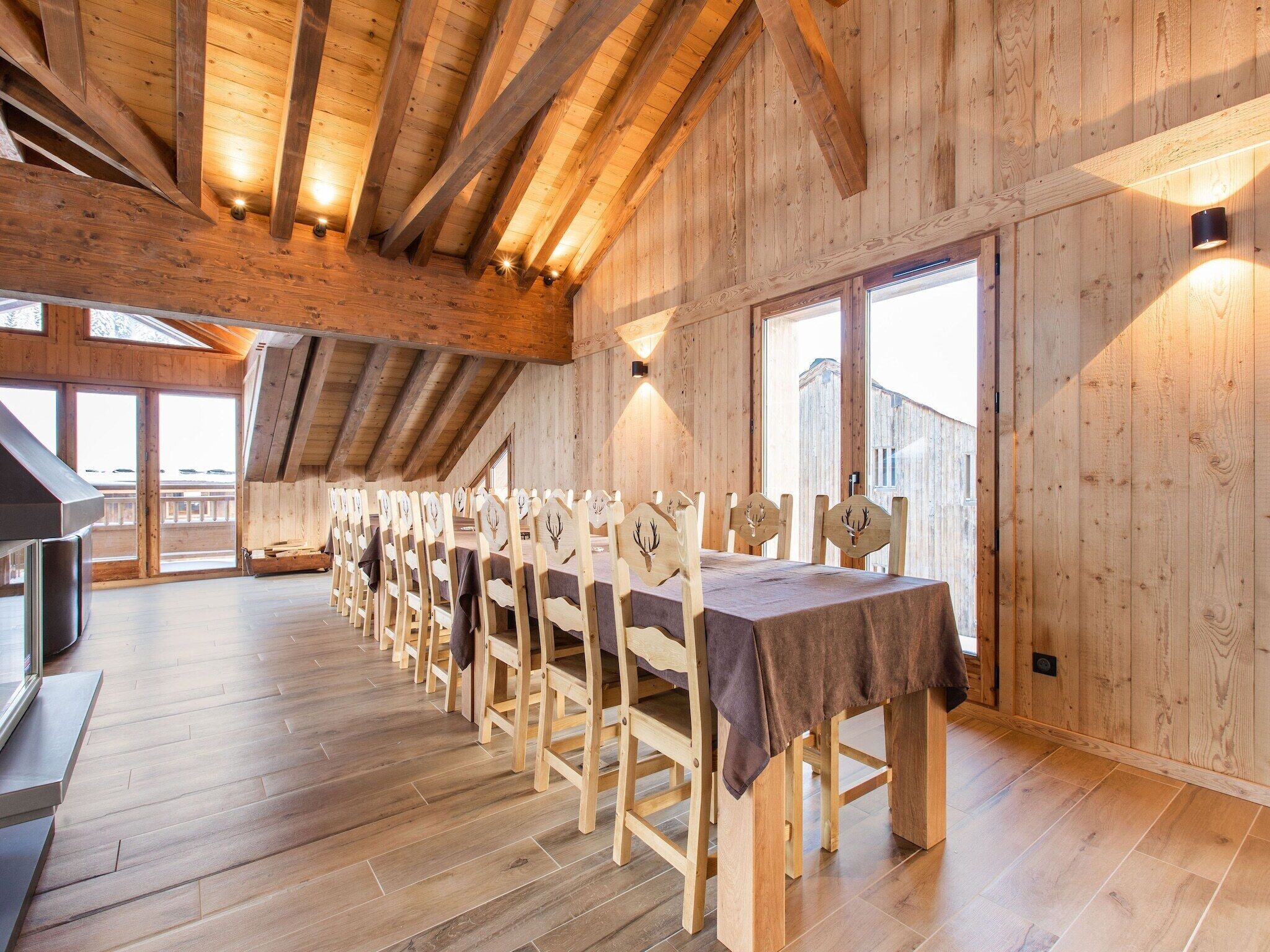 Une longue table à manger en bois, entourée de chaises, placée dans une pièce aux hauts plafonds, aux murs en bois et aux grandes fenêtres.