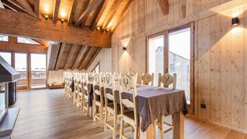 Une longue table à manger en bois, entourée de chaises, placée dans une pièce aux hauts plafonds, aux murs en bois et aux grandes fenêtres.