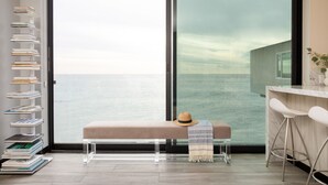 A modern room with a large window overlooking the sea, a bench with a hat and scarf, and a bar area with stools.