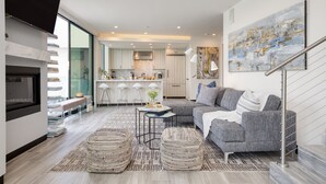 A modern living room with a grey sofa, a coffee table, and wicker ottomans. There is a kitchen in the background with bar stools and a large painting on the wall.