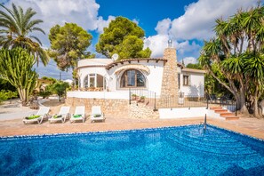 A poolside area with a clear blue pool, white lounge chairs, and a stone building with a tiled roof.