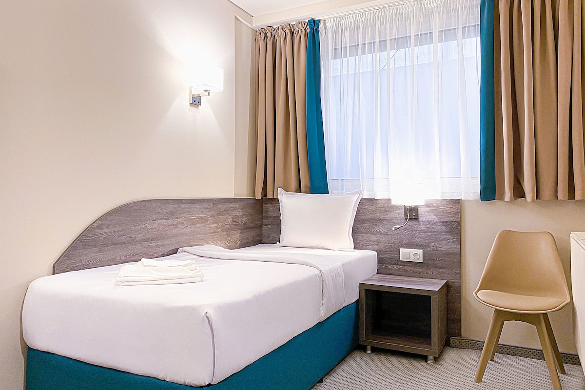 Quarto de hotel com cama, mesa de cabeceira, cadeira e janela com cortinas.