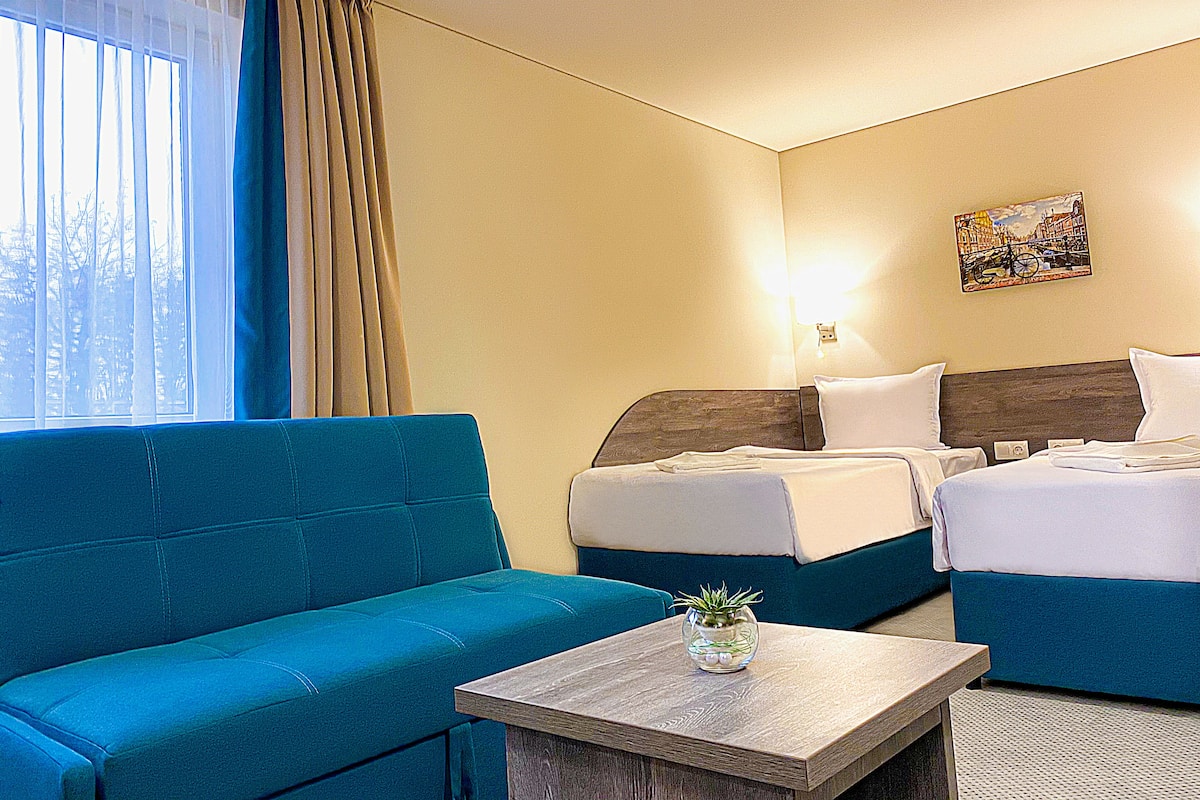 Quarto de hotel com sofá azul, duas camas de solteiro, uma mesa pequena com um vaso e um quadro na parede.