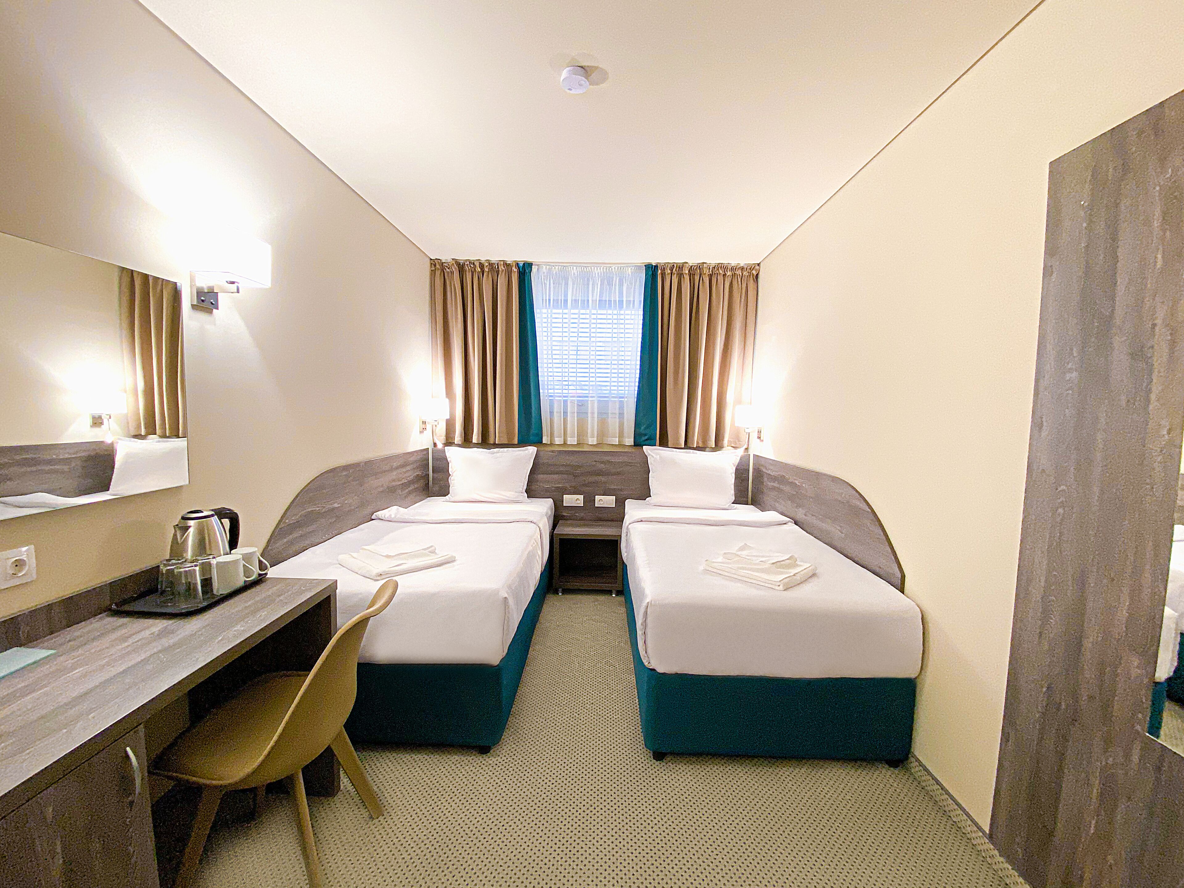 A hotel room with two beds, a desk, a chair, a kettle, and a window with curtains.