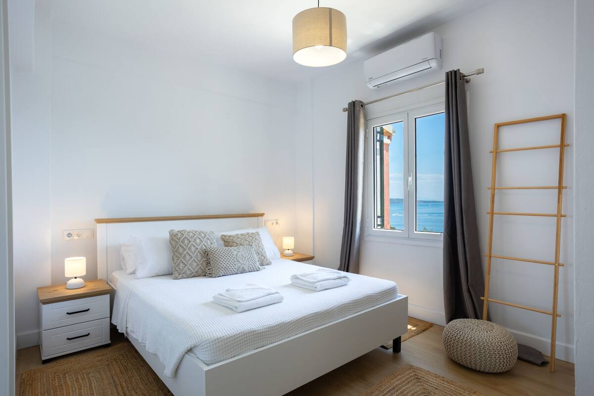 Ein ordentlich eingerichtetes Schlafzimmer mit einem Bett, Nachttischen, einer Lampe, einem Fenster mit Vorhängen und einer Holzleiter.