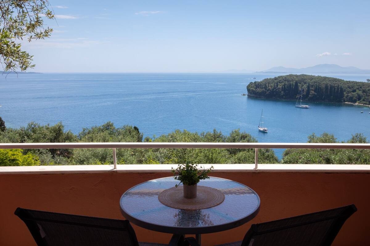 Ein Balkon mit rundem Tisch und Stühlen, der Blick auf ein ruhiges Meer mit Booten und einer bewaldeten Insel freigibt.